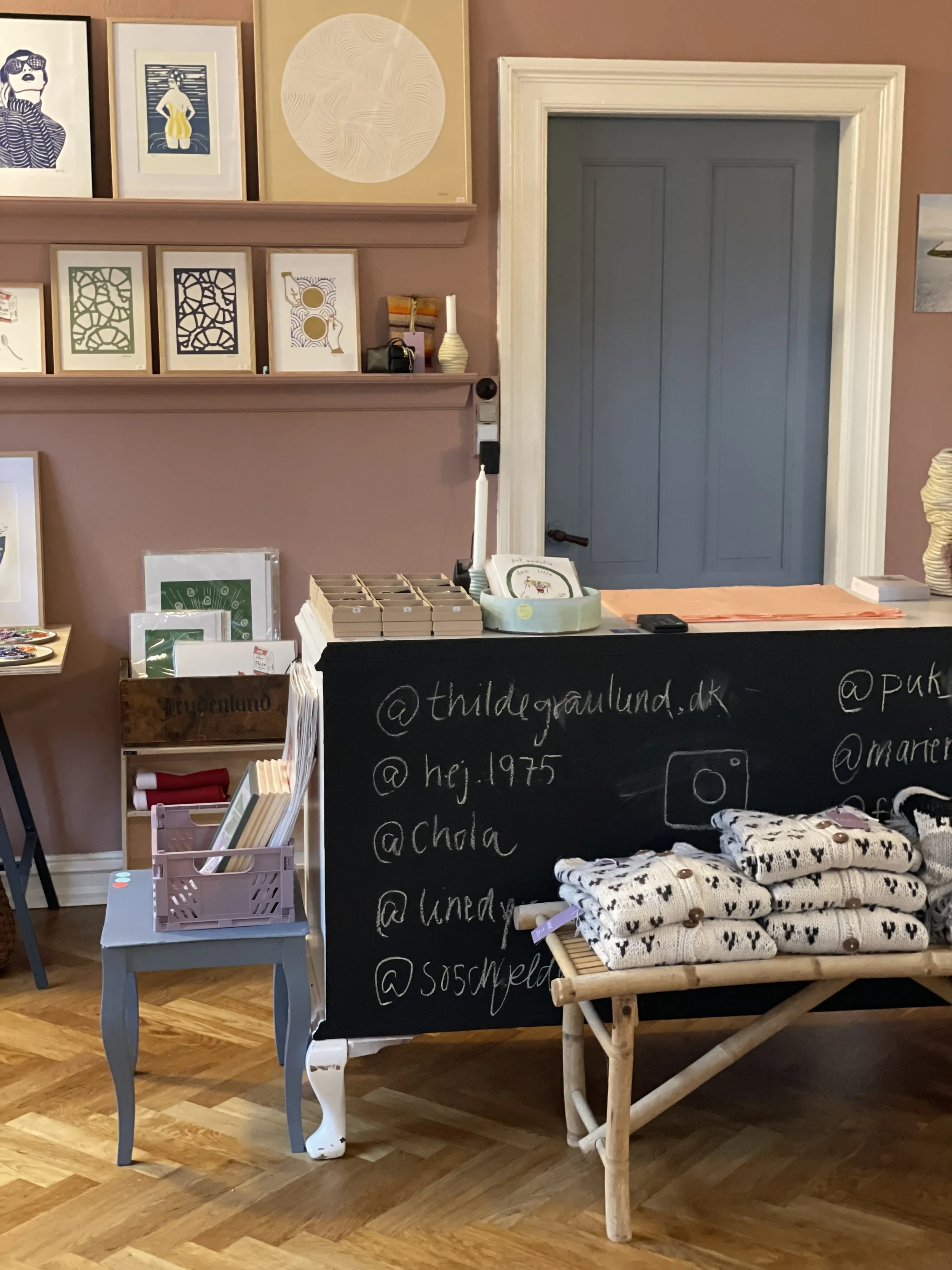 Indretning af en butik med hylder med kunstværker på væggen, en sort tavle med sociale mediehåndtag, en bænke med strikkede produkter, og en lille håndlavet træbord.