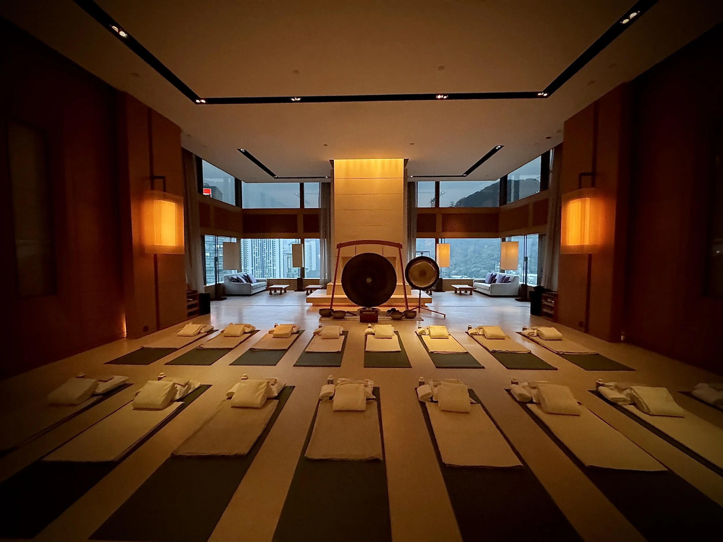 A dimly lit room set up for a meditation or yoga class, with low tables and folded towels or cushions on them, large gong and drum in the center, and floor-to-ceiling windows with a city and mountain view.