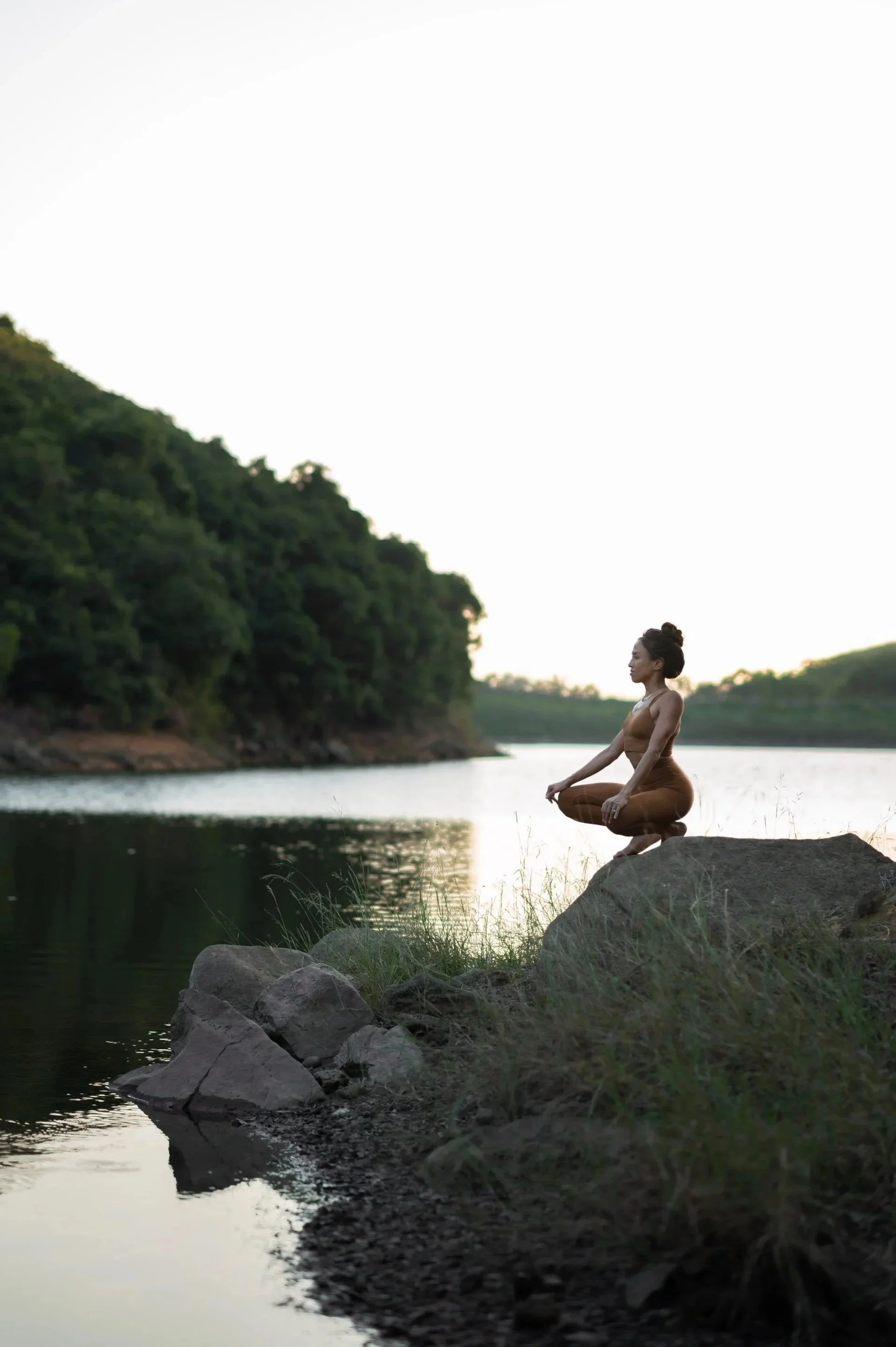 A woman practicing yoga or meditation on rocks by a calm river during sunset or sunrise, with lush green trees on the opposite riverbank.