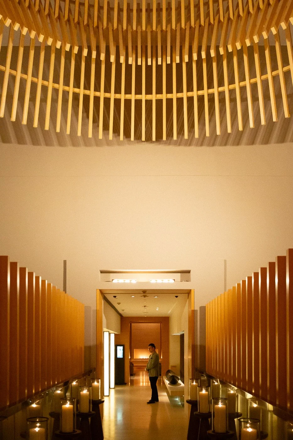 Interior of a modern building with a man standing in a corridor between wooden partition walls, with candles on tables along the sides and a wood ceiling feature above.