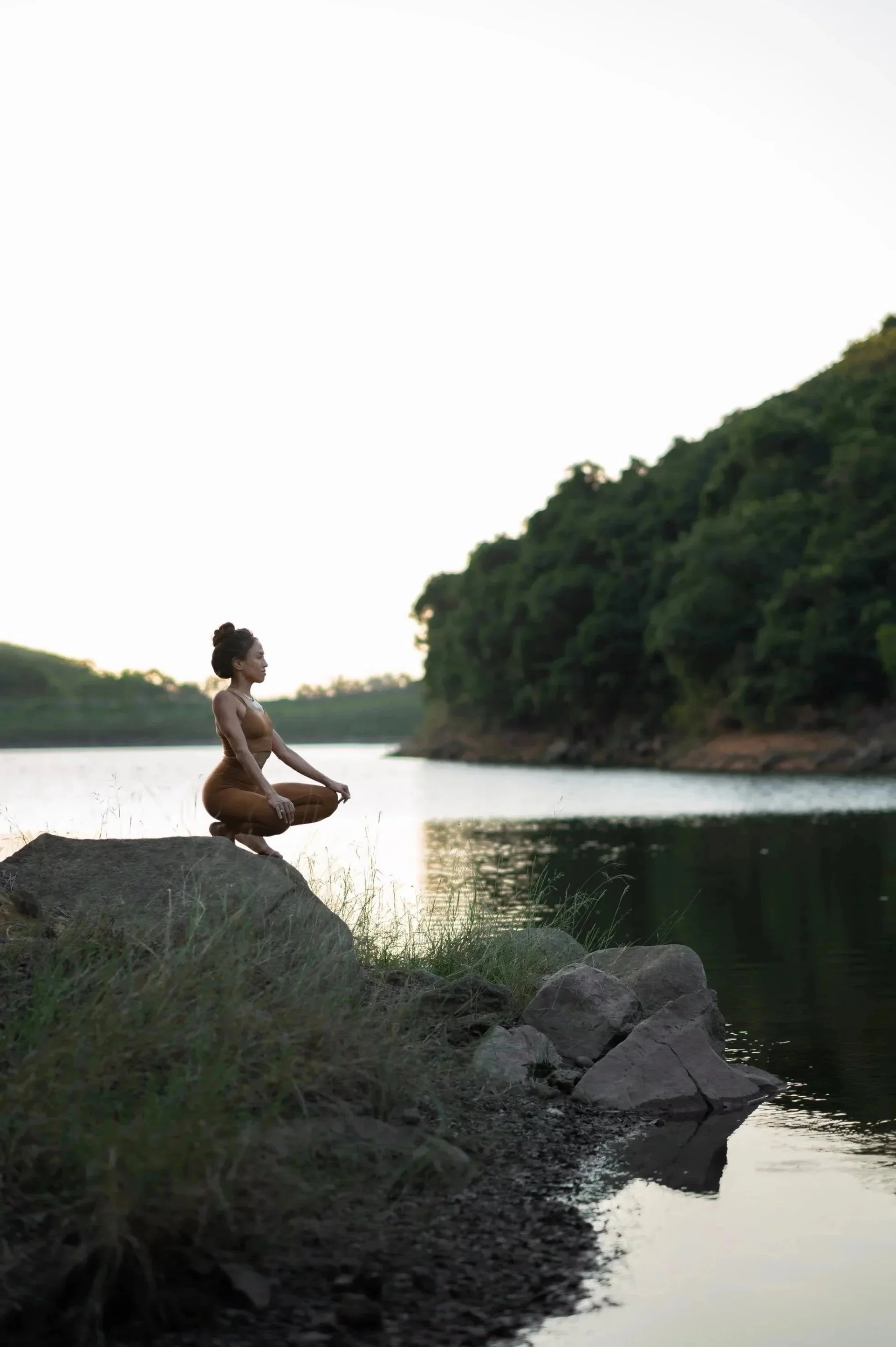 A woman in athletic wear sitting on a large rock by a peaceful river at sunset, surrounded by greenery and trees.