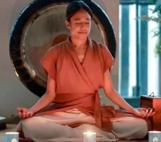 Woman meditating in a cross-legged seated position with candles in front, large gong in background, and natural light from window to the right.
