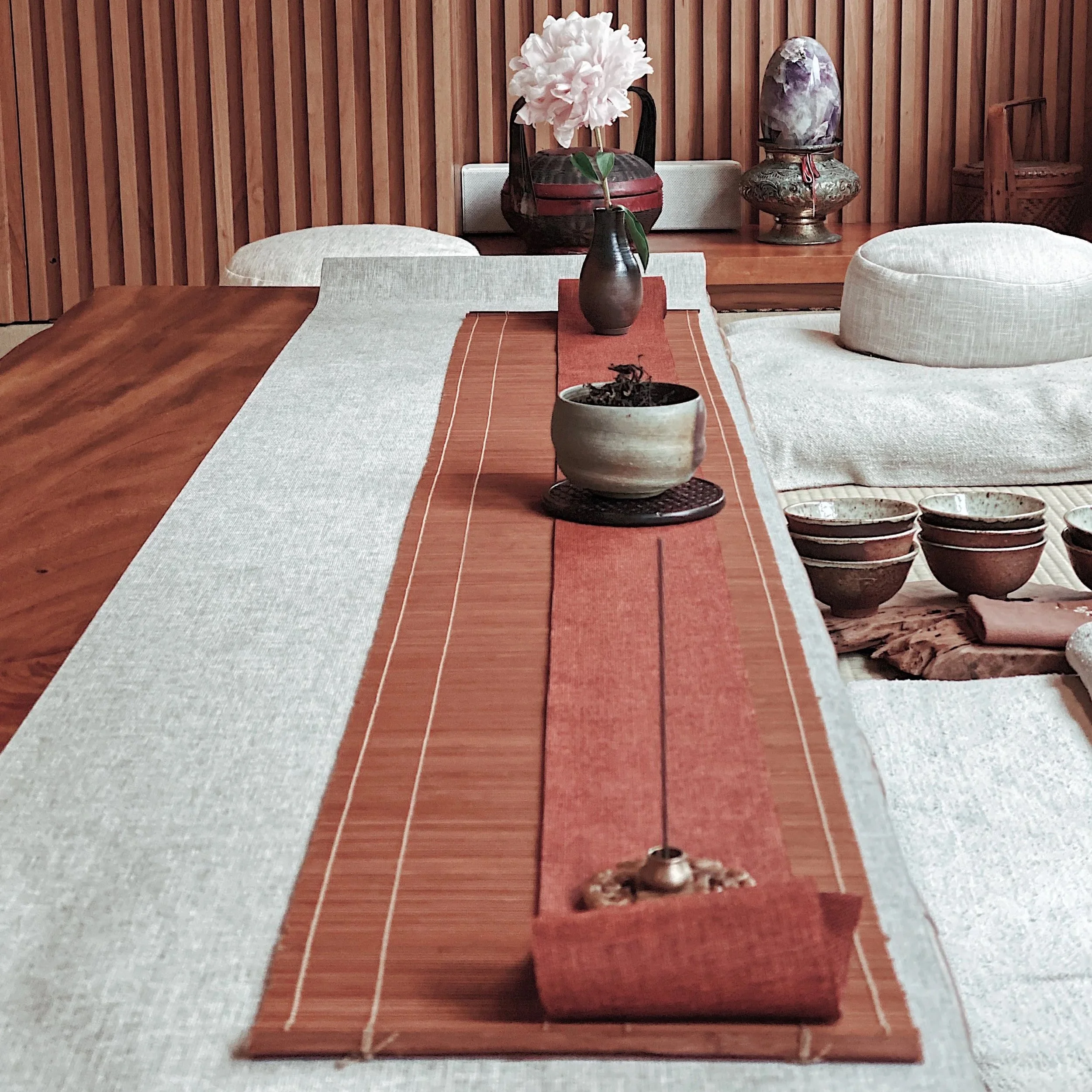 A wooden table with a beige table runner, decorated with a small potted plant and a small object, possibly a snack. In the background, a larger vase with a white flower, a decorative urn with purple and green patterns, and a wooden chair are visible against a wooden slat wall.