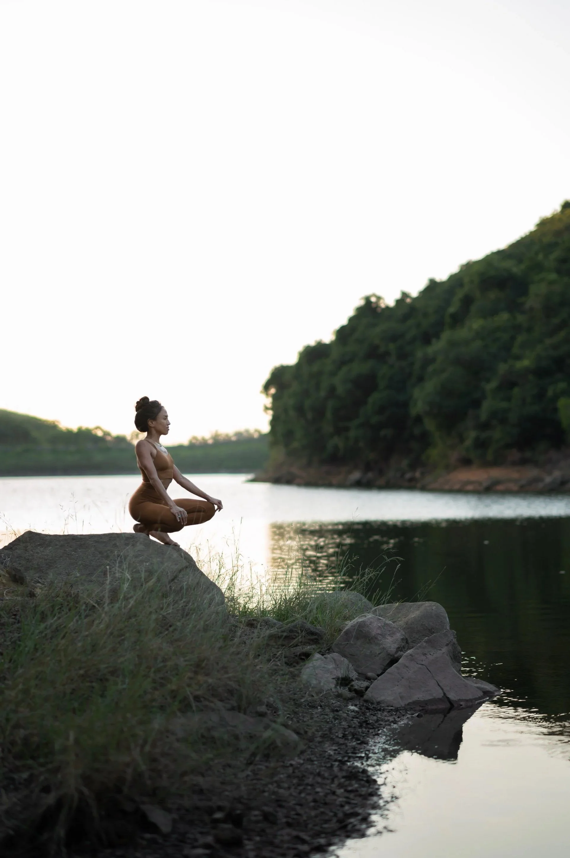 A woman practicing yoga outdoors by a river at sunset, sitting on a rock with her legs crossed and hands resting on her knees, surrounded by trees and rocks.