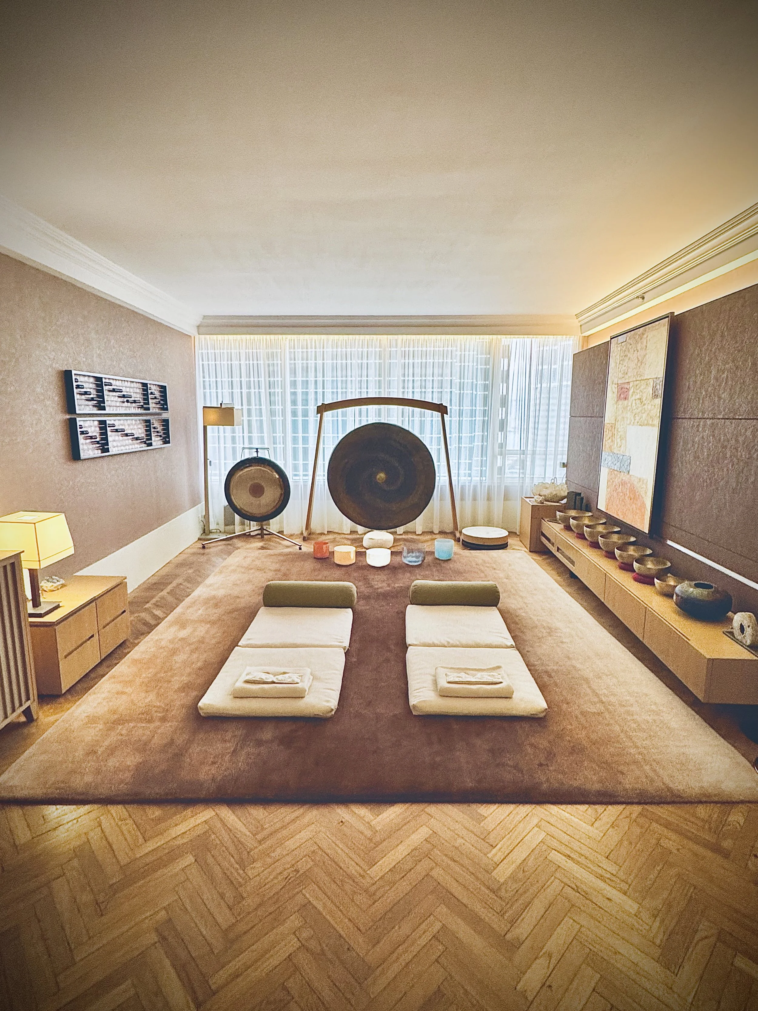 A room with a large brown carpet, two white padded mats with folded towels on top, a collection of singing bowls, gongs, and meditation cushions, and soft lighting from windows with sheer curtains.