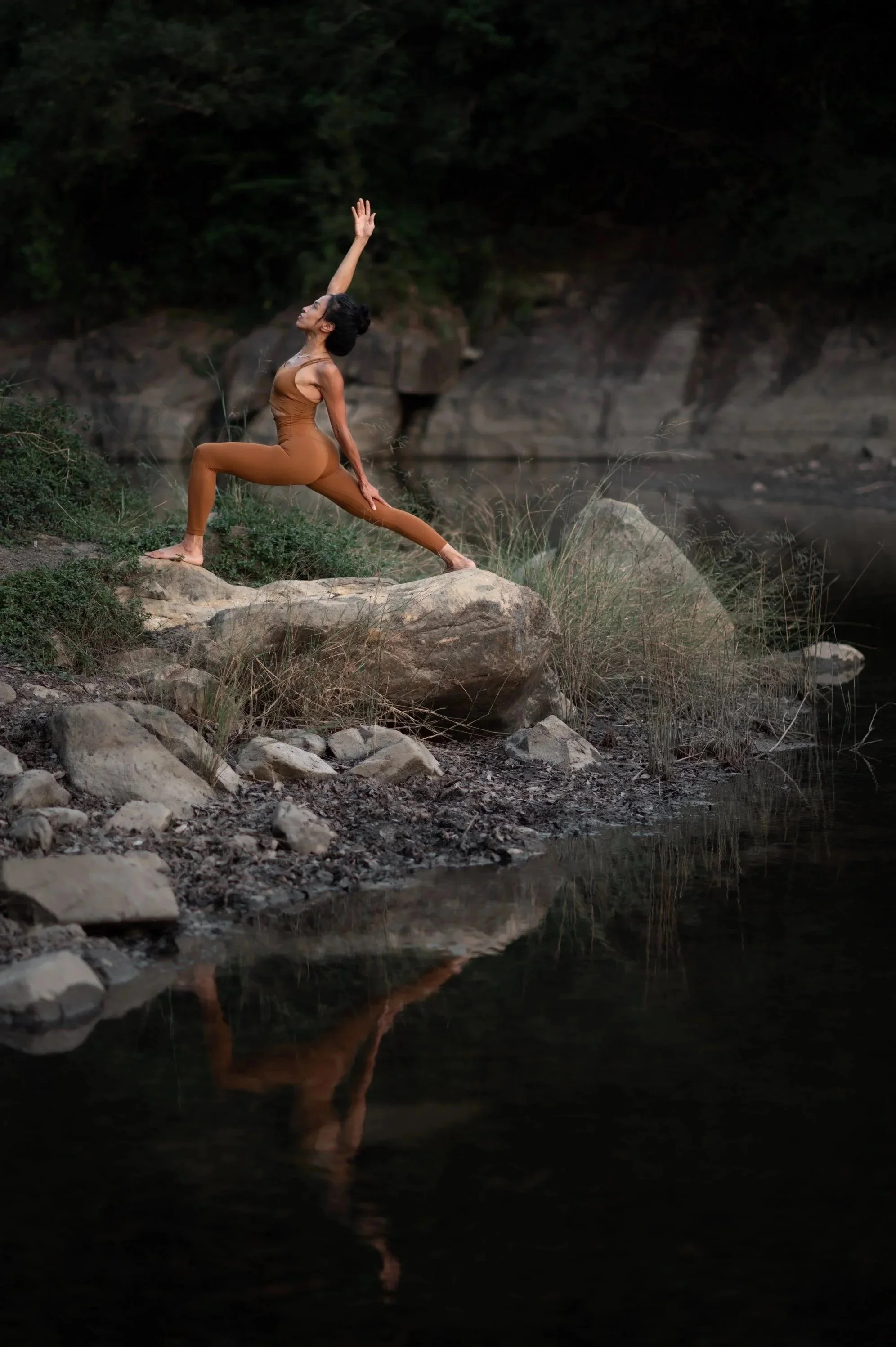 A woman practicing yoga in a lunge pose on rocks near a river at sunset.