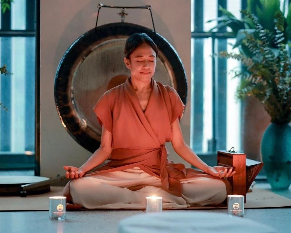 A woman meditating in a cross-legged seated position, with her eyes closed and fingers resting on her knees, in a room with candles and plants, with a gong behind her.