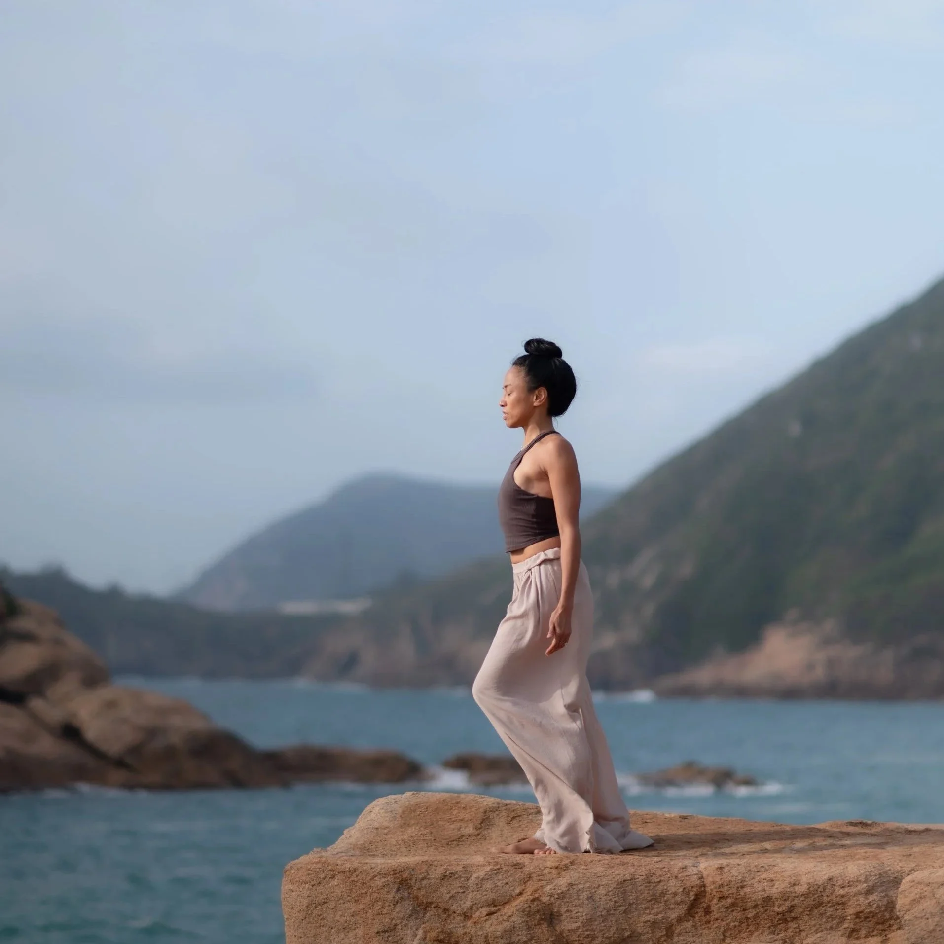 A woman practicing yoga outdoors on a rocky ledge near the ocean, with mountains and cloudy sky in the background.