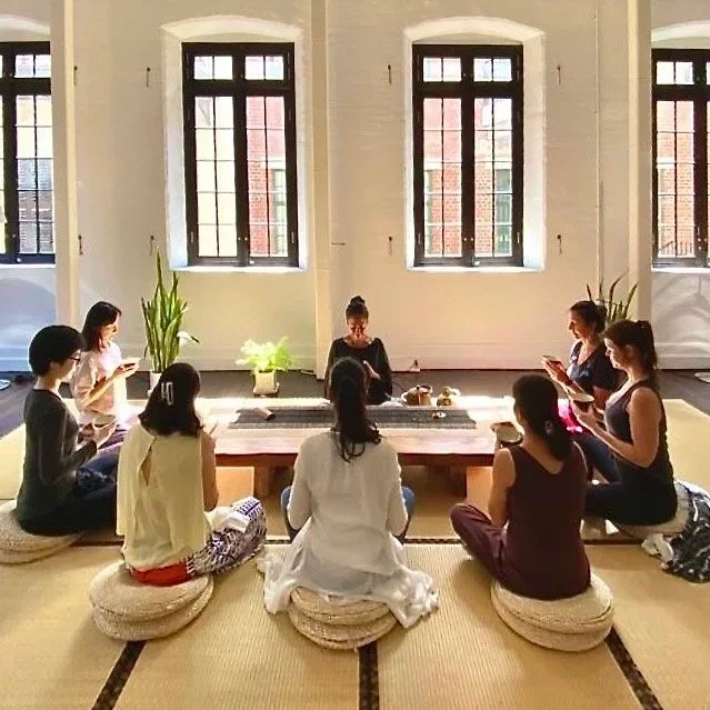 Ten women sitting in a circle on cushions in a bright room with large windows, some using their phones and one in the center with eyes closed, possibly meditating or leading a session.