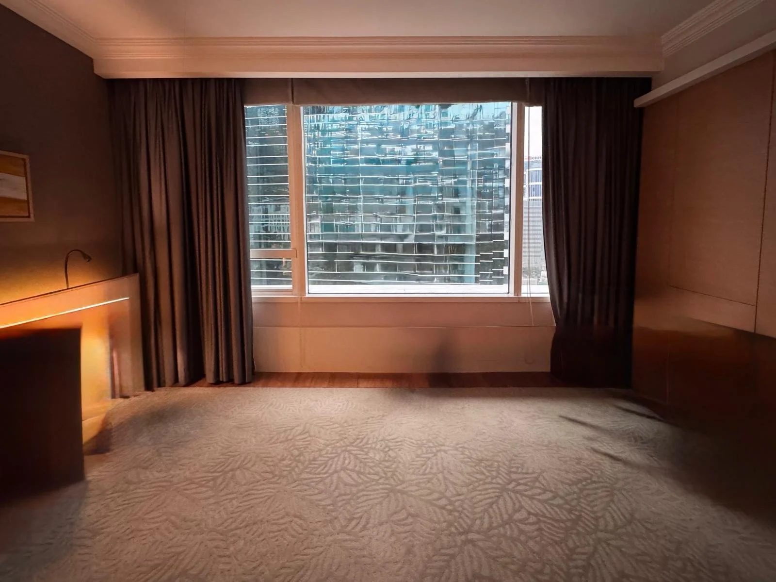 Empty hotel room with beige carpet, wooden furniture, dark curtains, and large window with view of neighboring modern glass buildings.