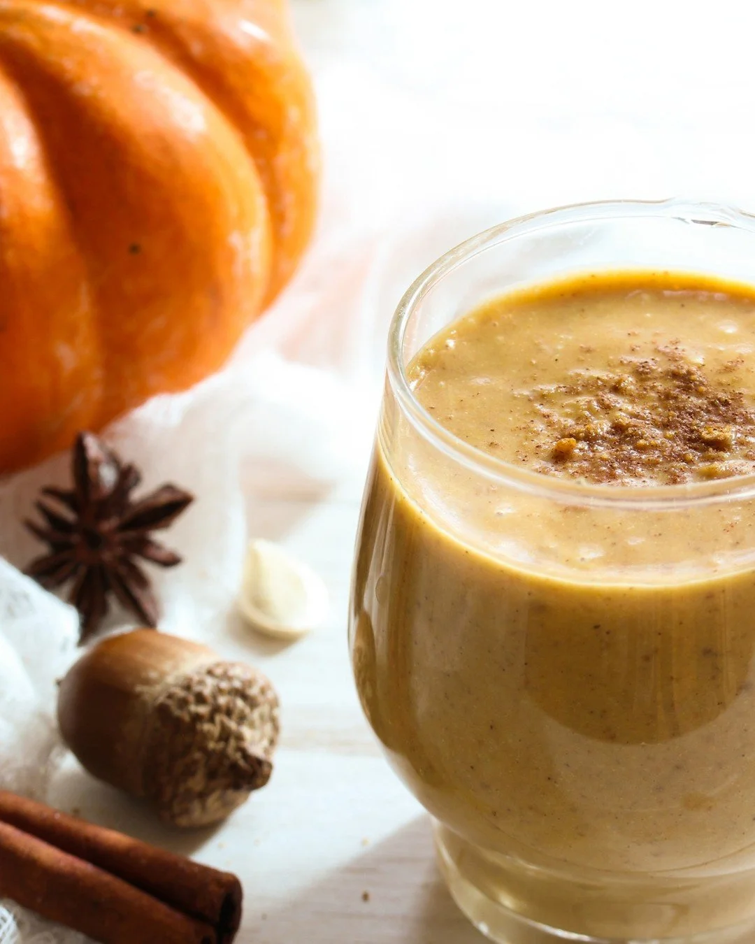 Pumpkin cheesecake protein smoothie in a glass with a sprinkle of spice, next to a small pumpkin and cinnamon sticks