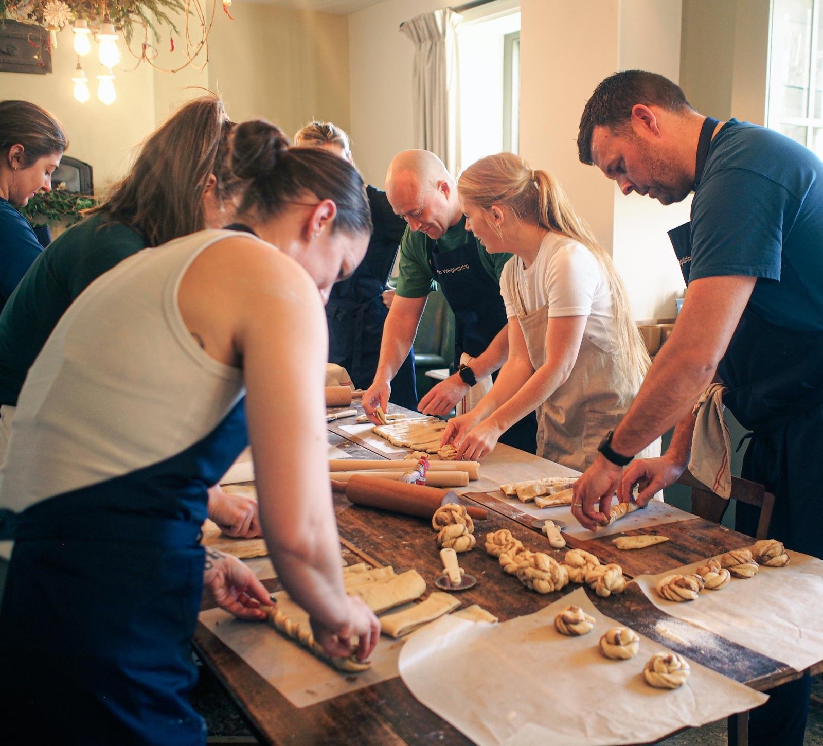 Group of people preparing and baking cinnamon rolls in a home kitchen. They are wearing aprons and working on a wooden table with rolling pins and dough.