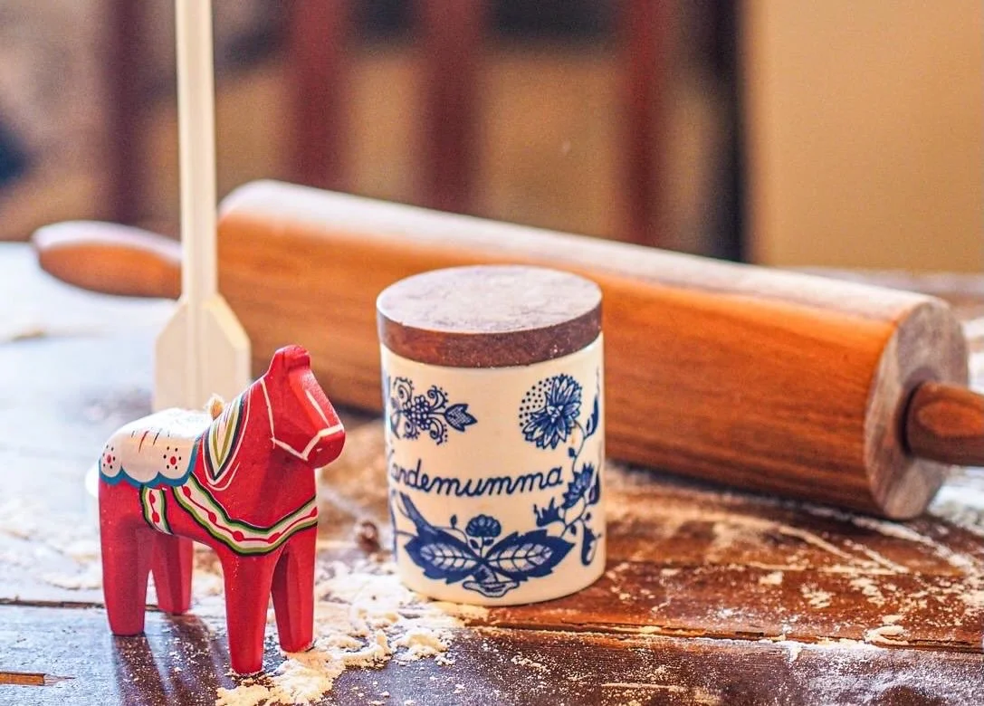 A wooden rolling pin, a decorative red and white horse, and a blue floral container with the word 'mormor' are on a floured wooden surface.