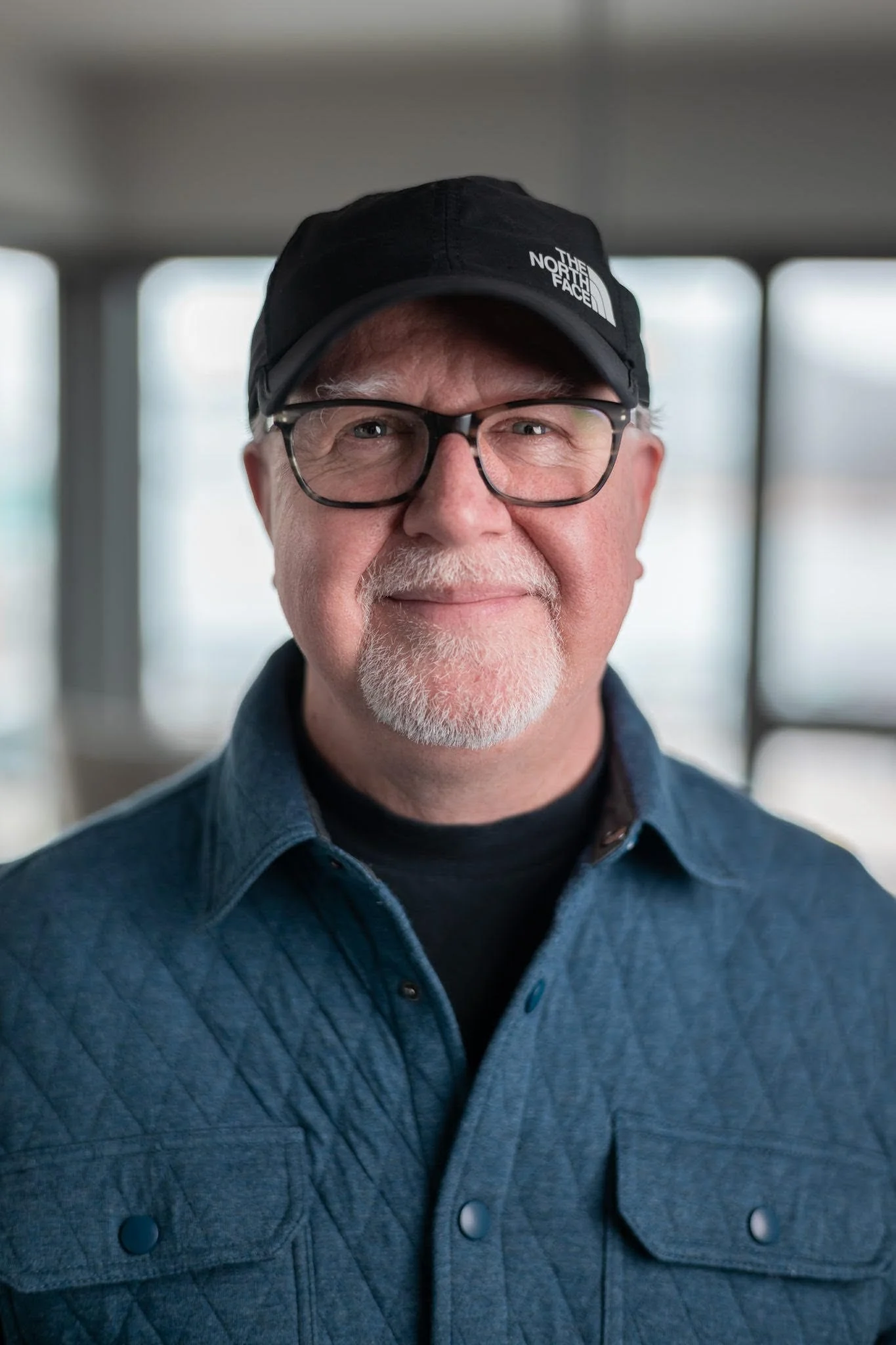 A middle-aged man with glasses and a goatee, wearing a black North Face cap and a blue button-up shirt, smiling at the camera indoors with blurred windows in the background.