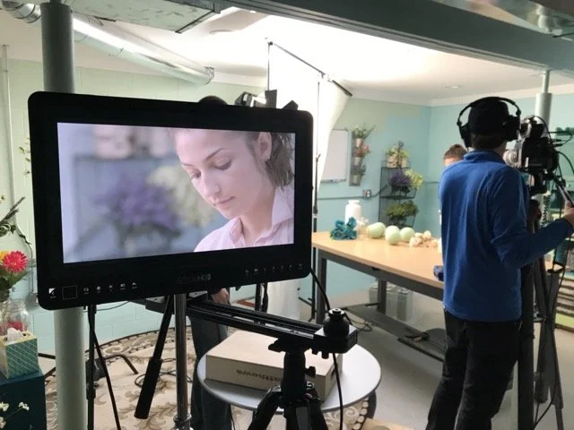 A woman on camera is being filmed in a studio with a man recording in the background. The studio has a blue wall, shelves with plants, and various props on a table.