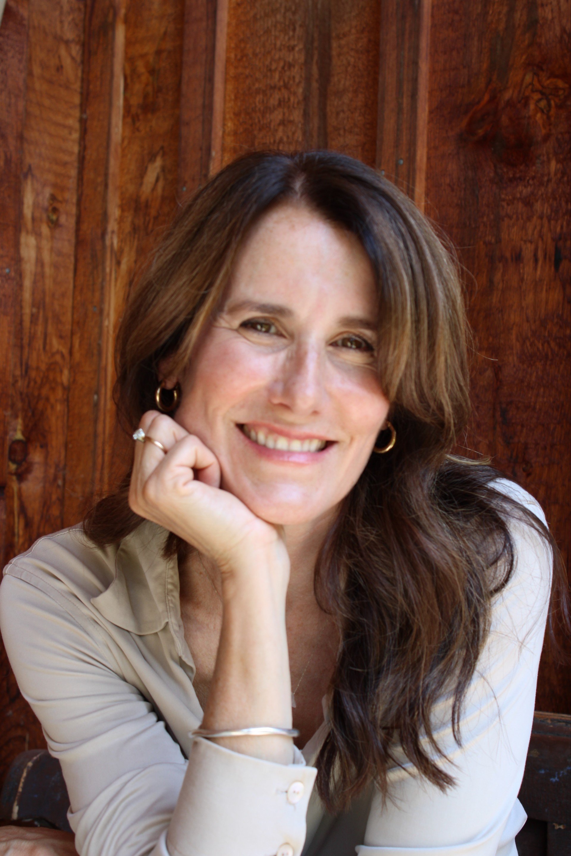A woman with long brown hair smiling, resting her chin on her hand, sitting in front of a wooden background.
