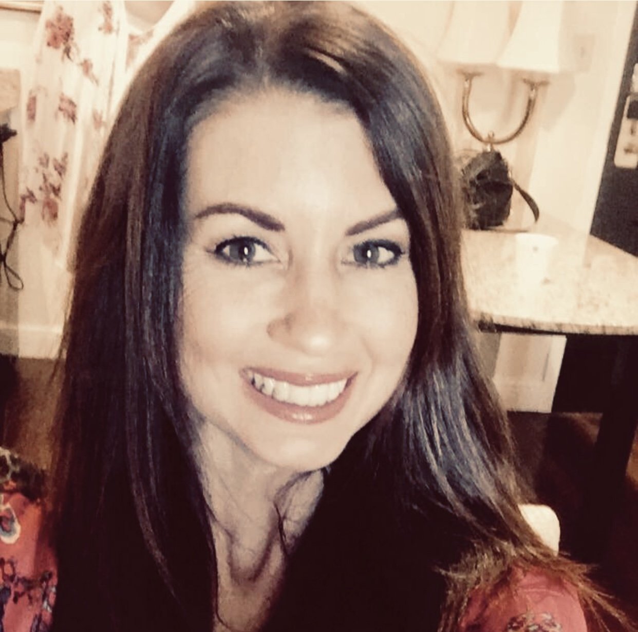 Close-up of a smiling woman with long brown hair in a cozy indoor setting.
