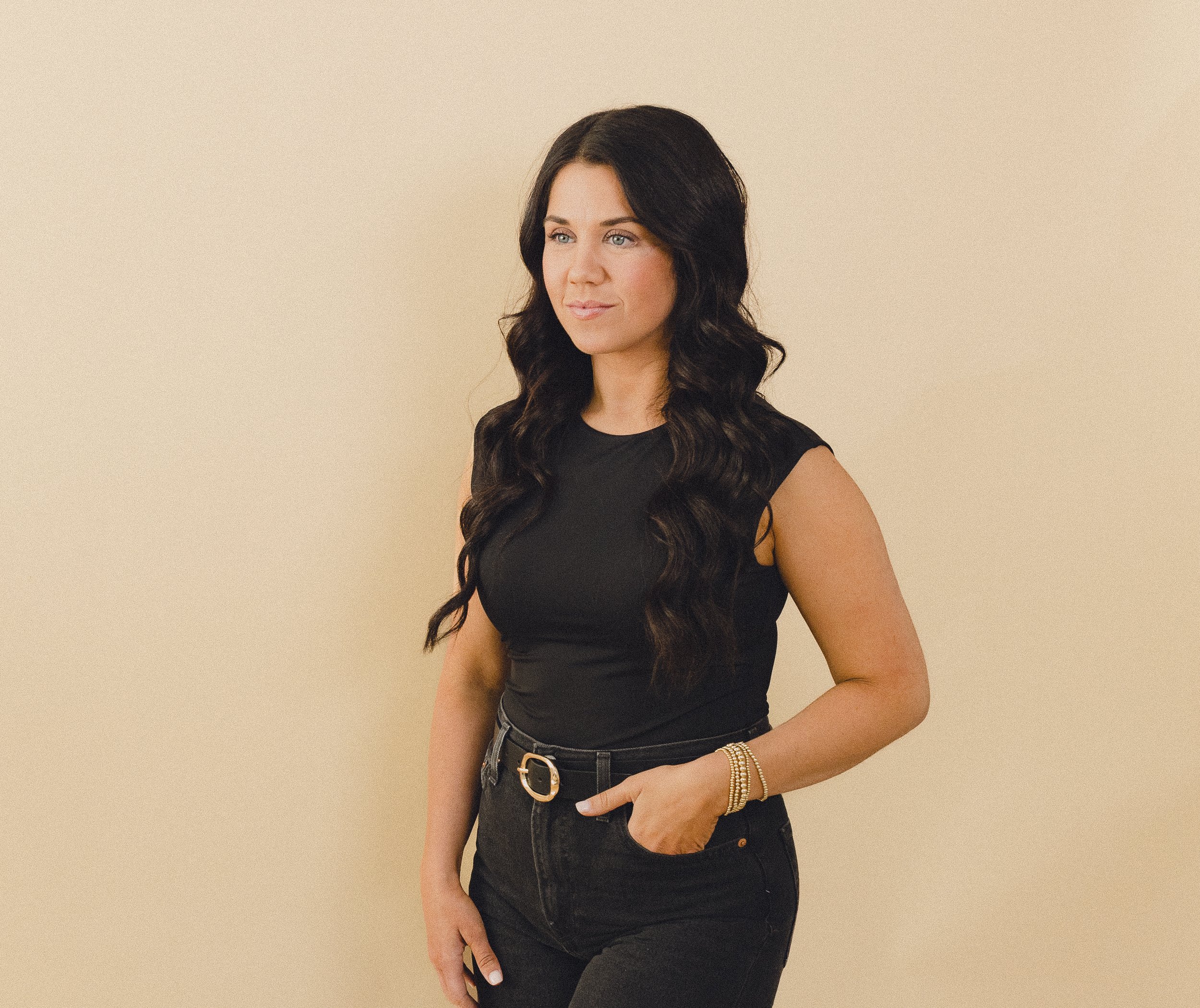 A woman with long, dark, wavy hair wearing a black sleeveless top and black jeans, standing against a plain beige background.