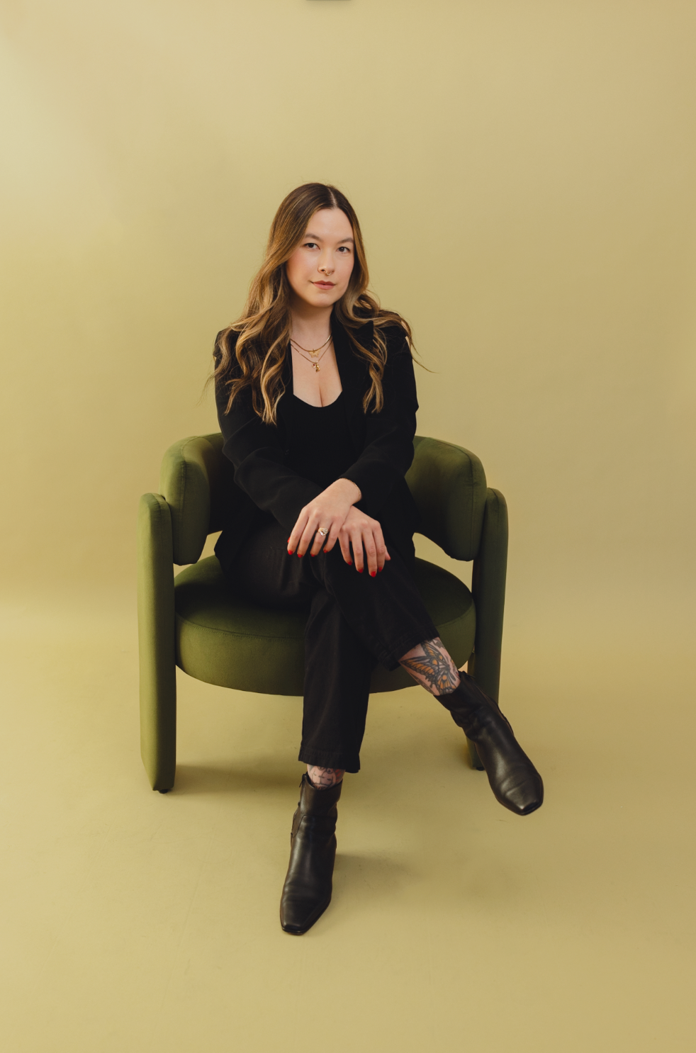 Permanent Makeup Artist Emily with long, wavy hair, sitting on a green armchair against a plain, light-colored background, dressed in black clothing with jewelry and black boots.