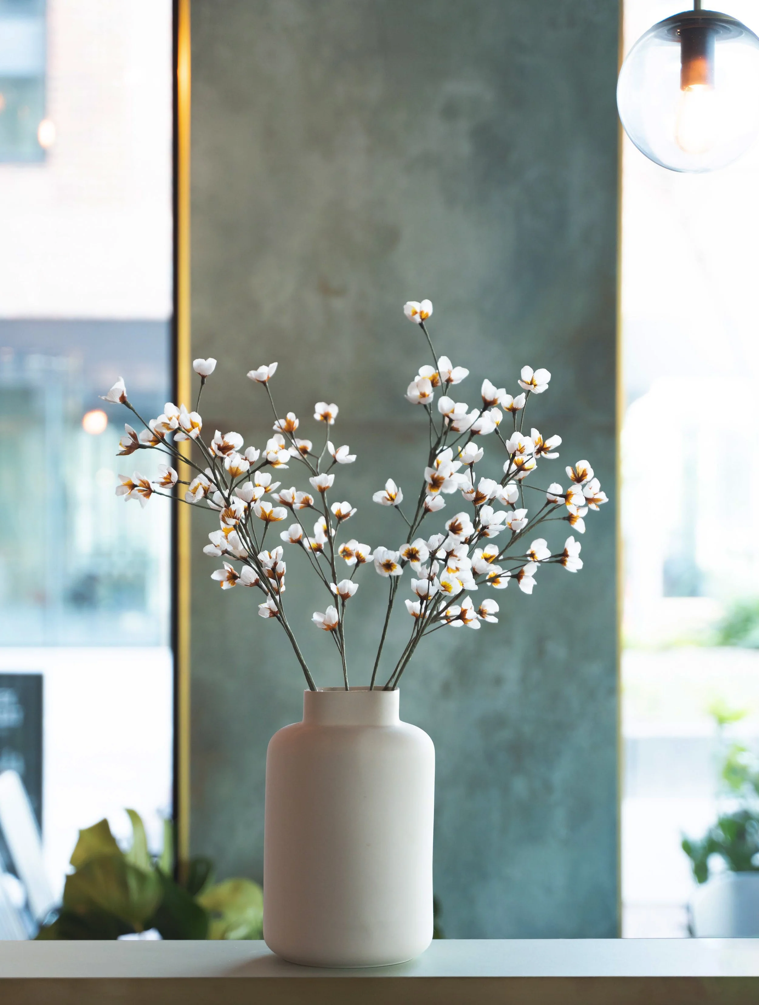 En hvid vase med hvide blomster lavet af papir på en hvid overflade indendørs.