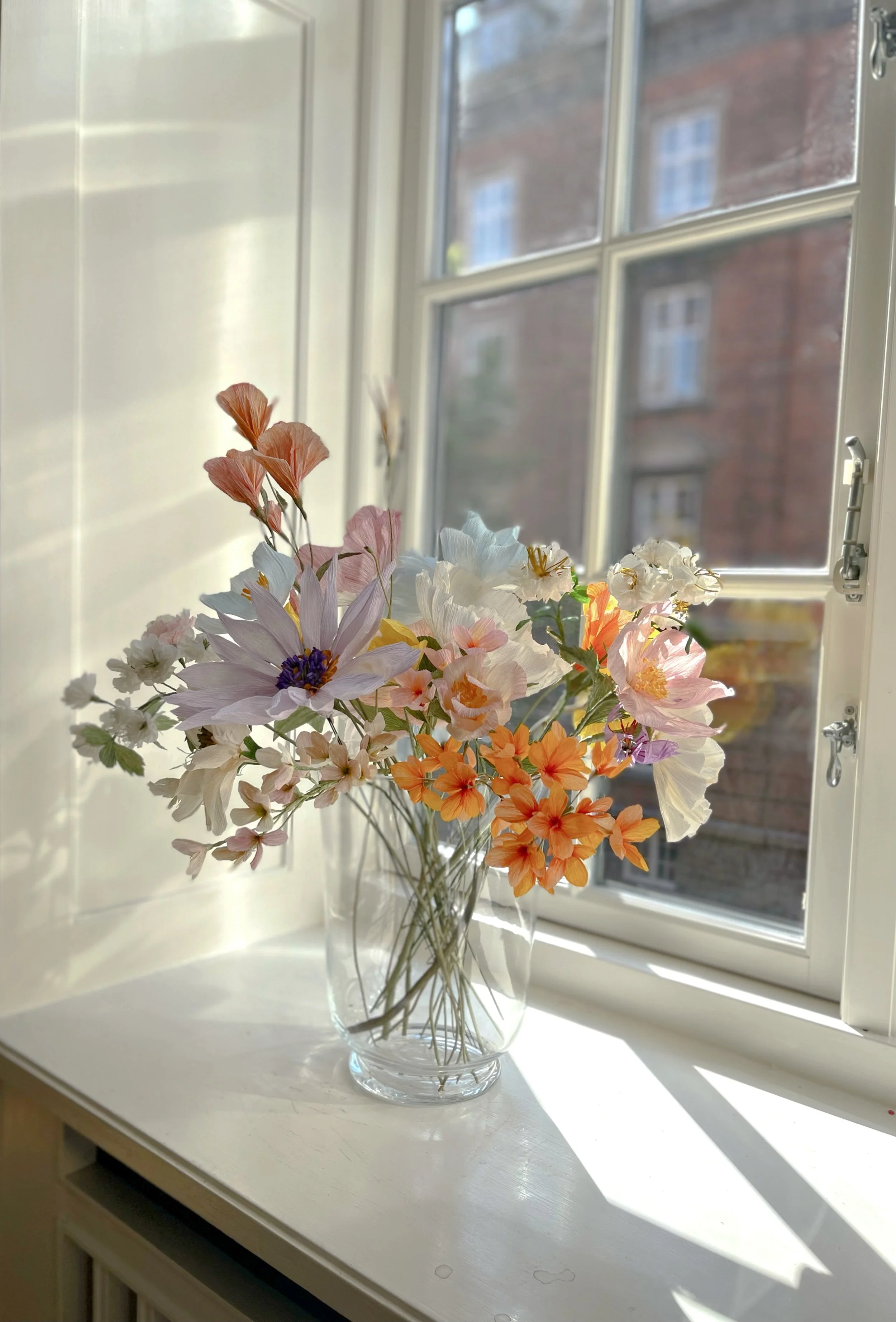 En gennemsigtig vase med lyserøde, orange, tofargede og hvide blomster står på en hvid vindueskarm, med sollys der skinner gennem vinduet.