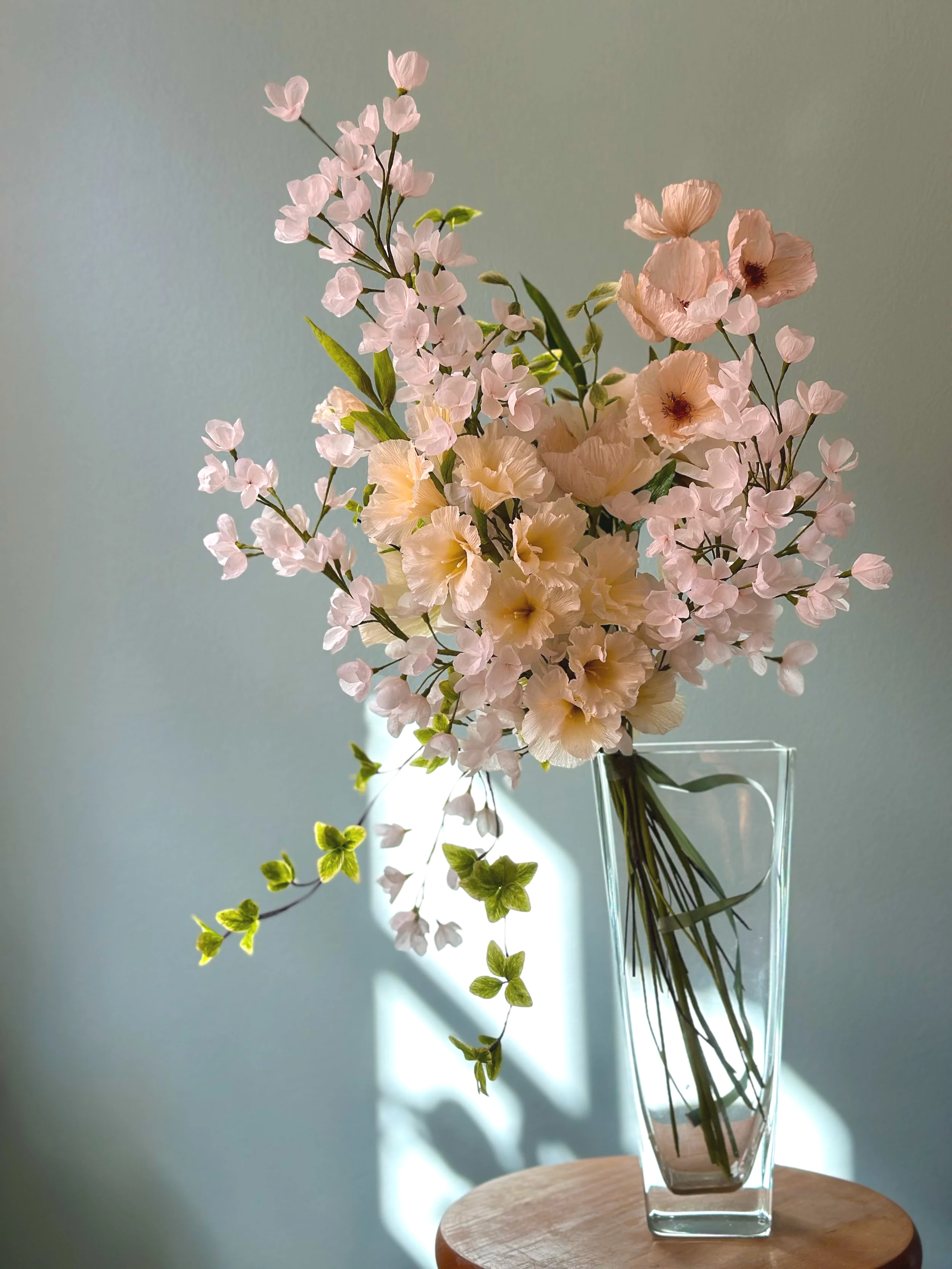 Lyseblomstret buket med lyserøde og cremefarvede blomster i en gennemsigtig glasvase på et rundt trækald.
