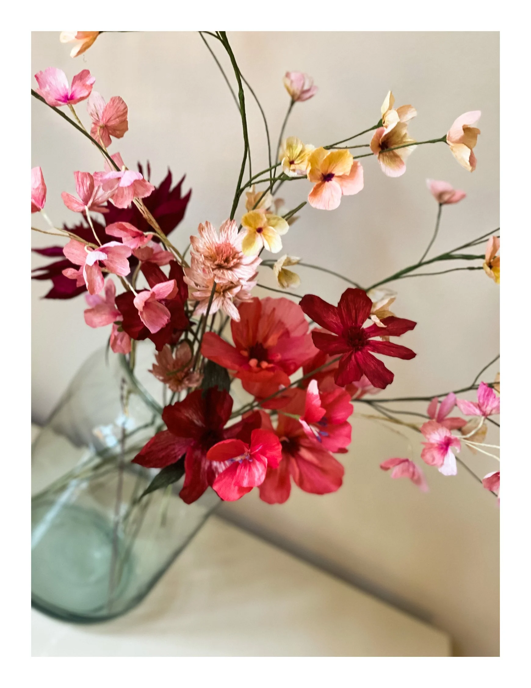 Buket af farverige blomster lavet af papir i en glasvase på en hvid overflade.