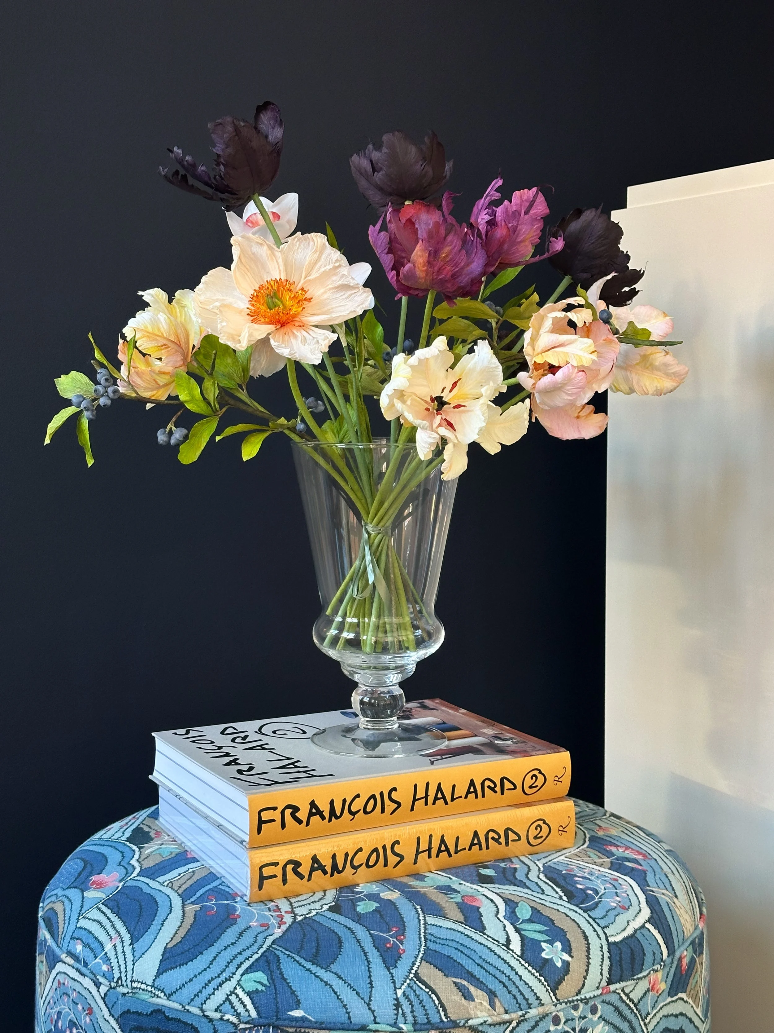 En glasvase med en blomsterbuket med lyserøde, lilla og mørke blomster, placeret på to bøger med gule og hvide omslag, som står på en blå, mønstret skammel.