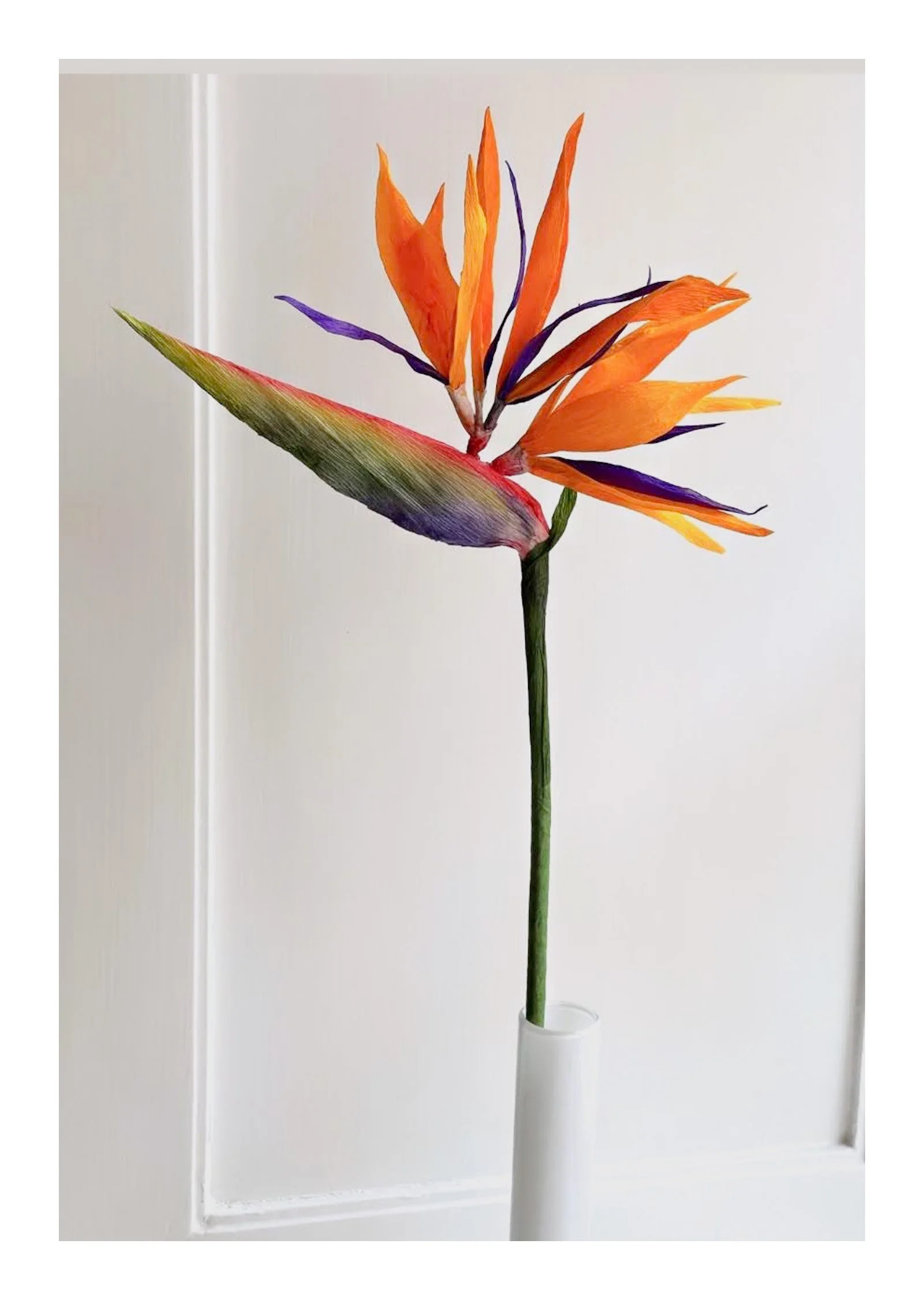 En kunstig blomsterdekoration med orange, lilla og grønne blade i en hvid vase, placeret foran en hvid væg.