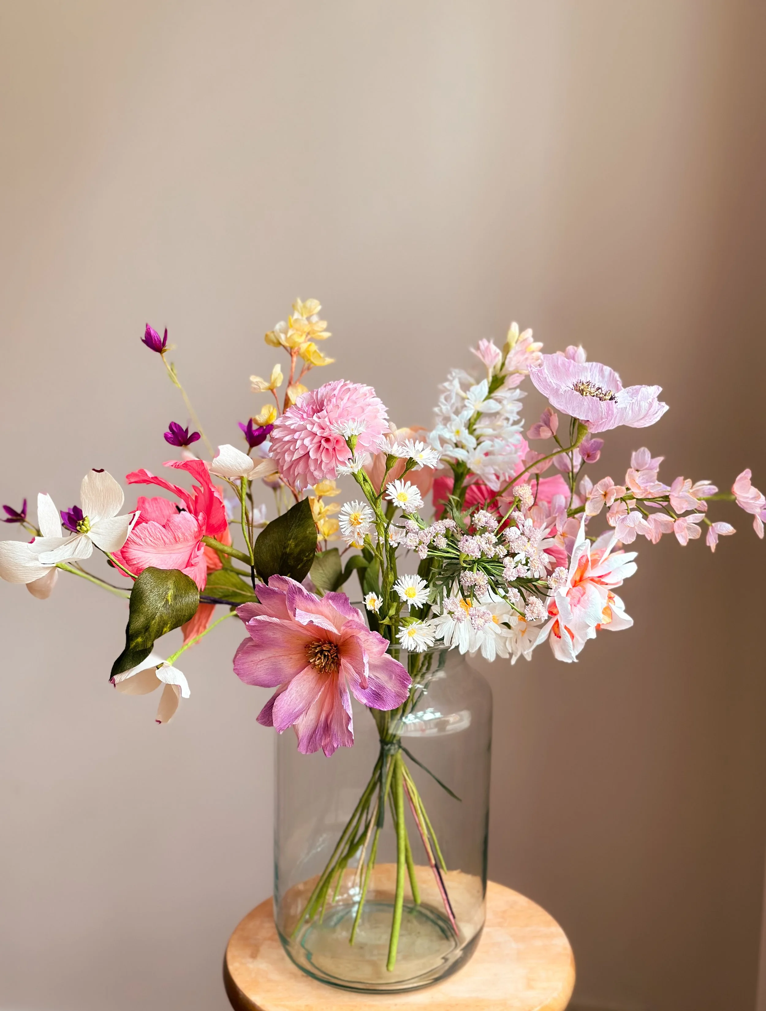 En gennemsigtig vase med en buket af forskellige lyserøde, hvide og lilla blomster, placeret på et rundt træbord mod en beige væg.