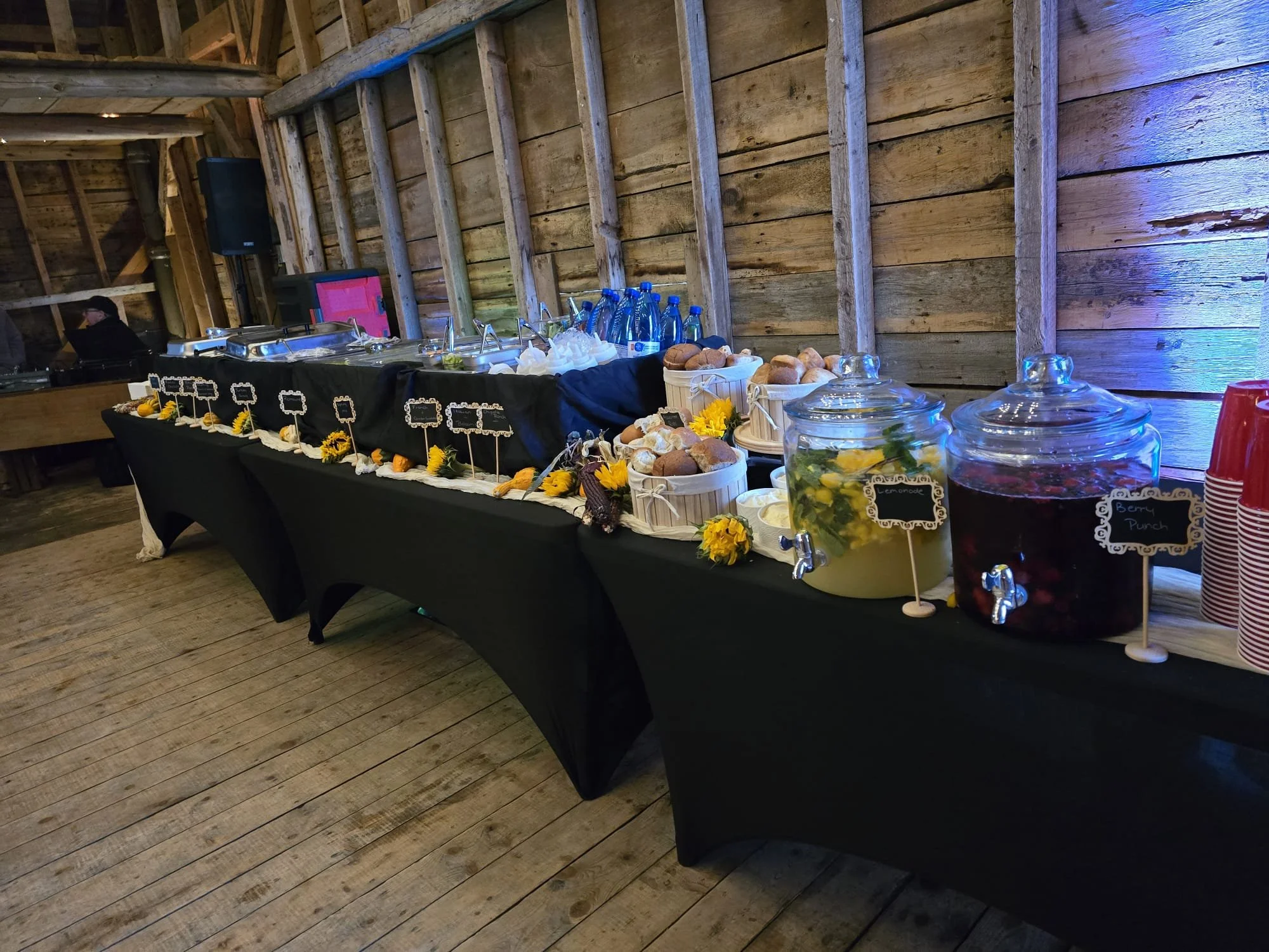 A buffet table of breads and other items, including cold drinks. The tables are covered in black tablecloths. The setting is the interior of a wooden barn.