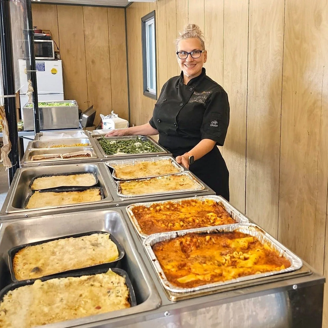 Chef in a kitchen with trays of prepared food, including lasagna and green beans, on stainless steel surfaces.