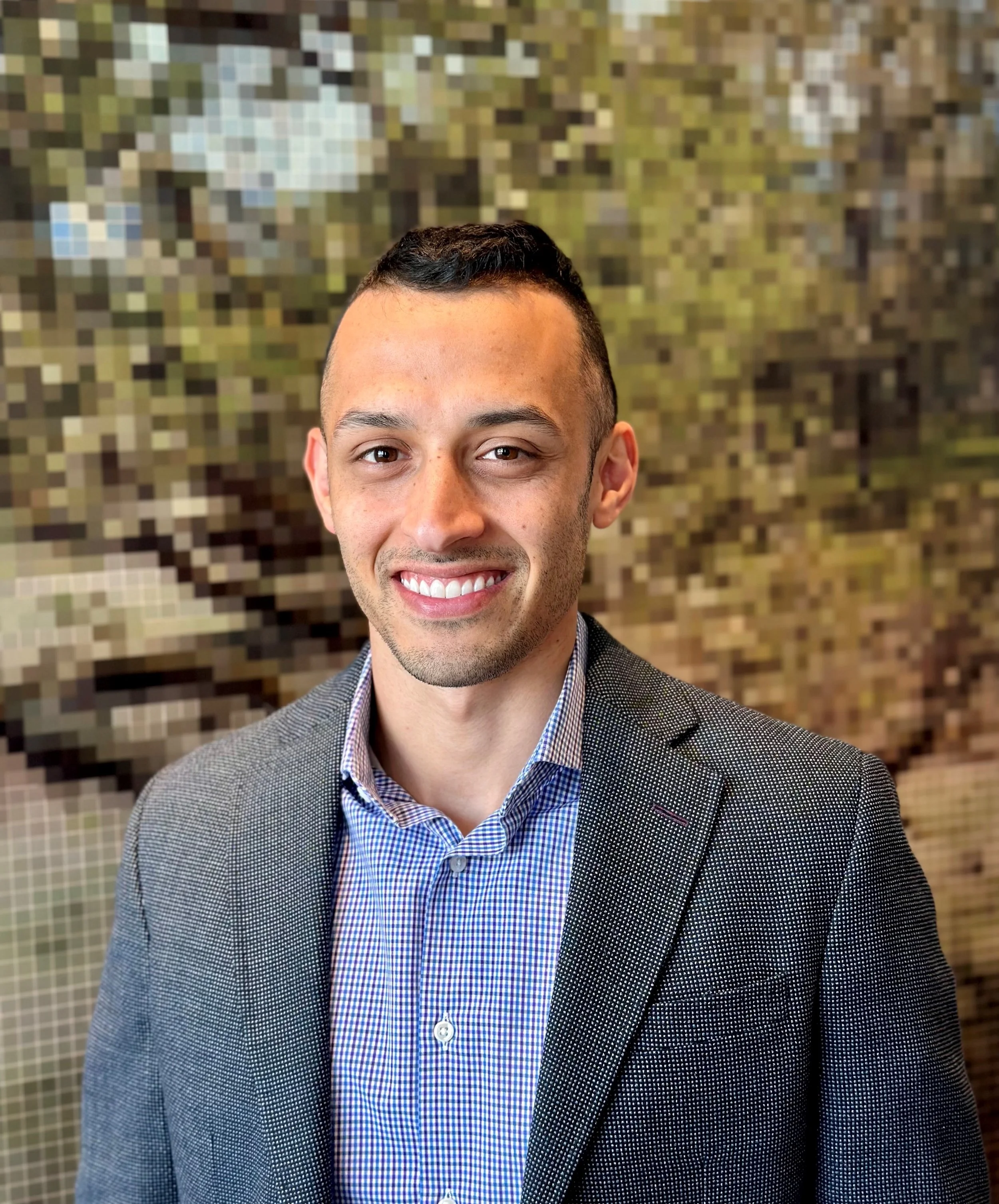 A man with short dark hair wears a button-up shirt and blazer