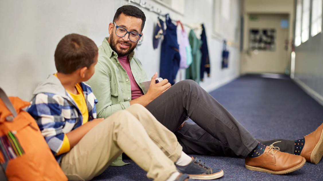 Adult sits on floor next to child and talks to them them
