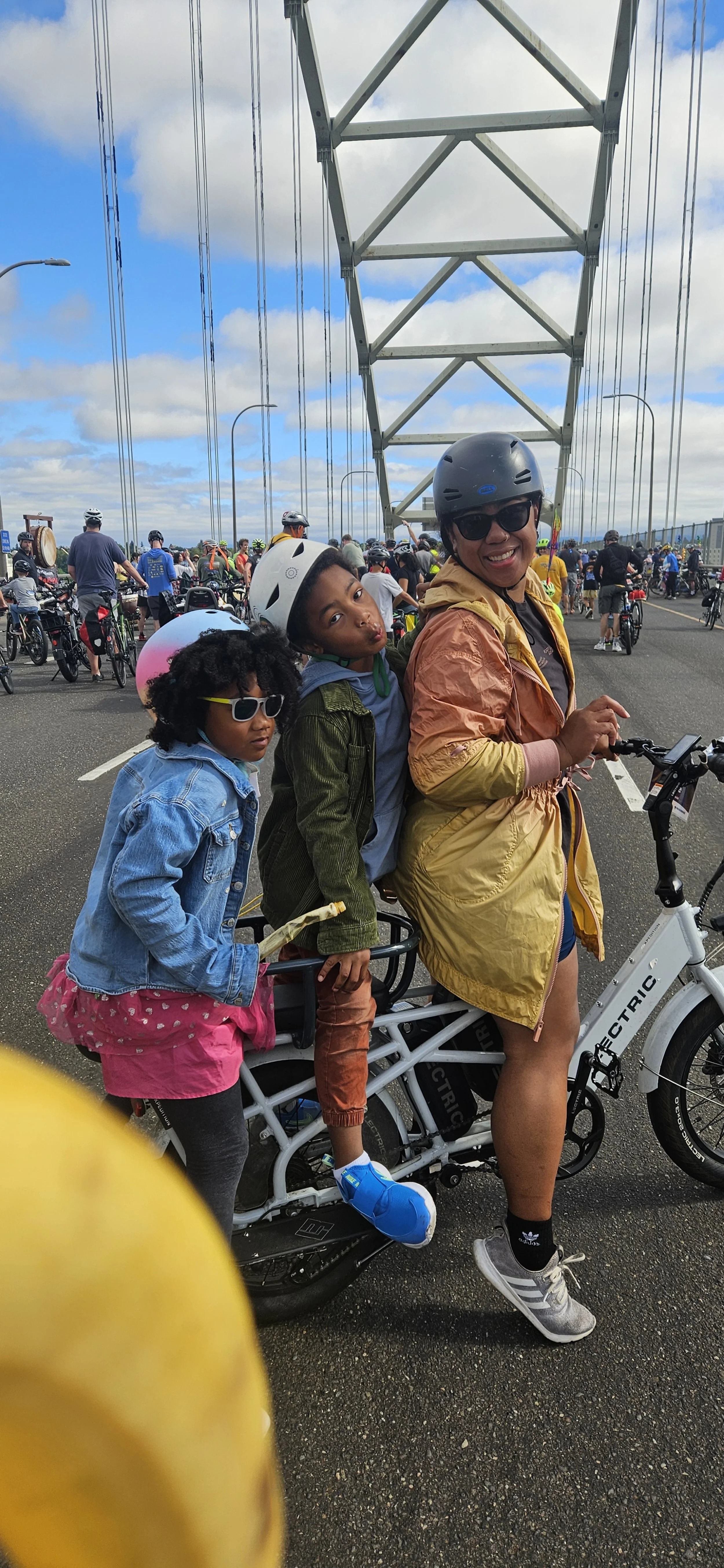 a woman on a cargo bike with two children wearing helmets and colorful outfits, all smiling.