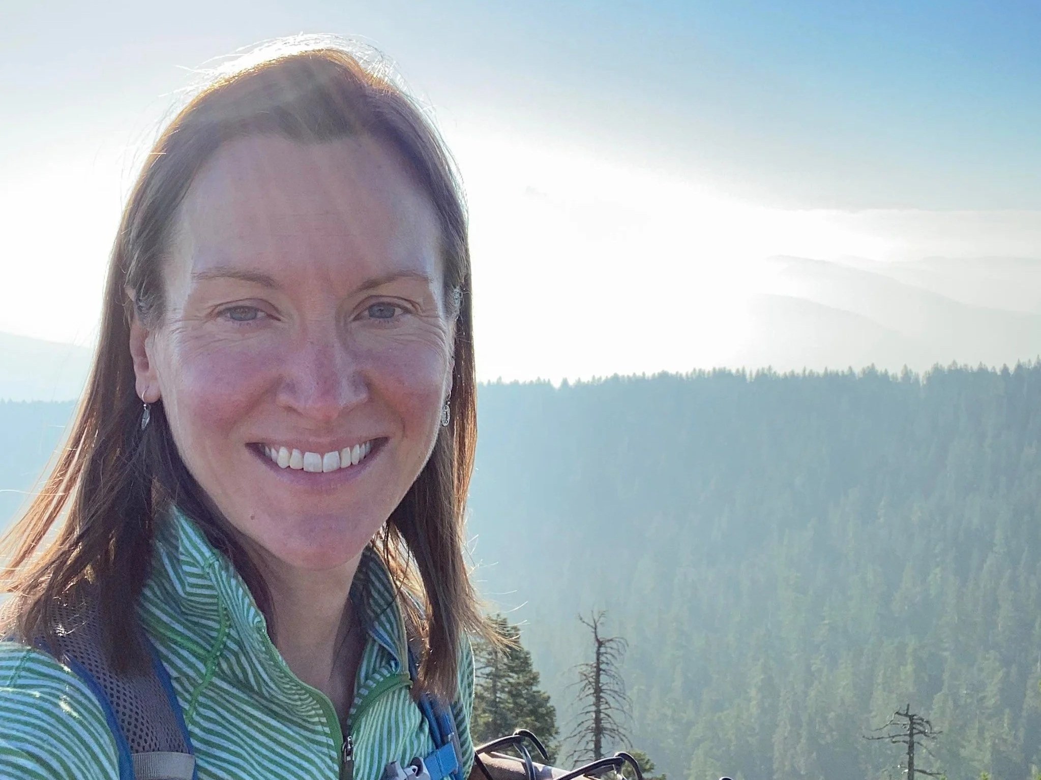 A woman with long light brown hair smiles outdoors on a sunny day atop a hill with trees in the background.