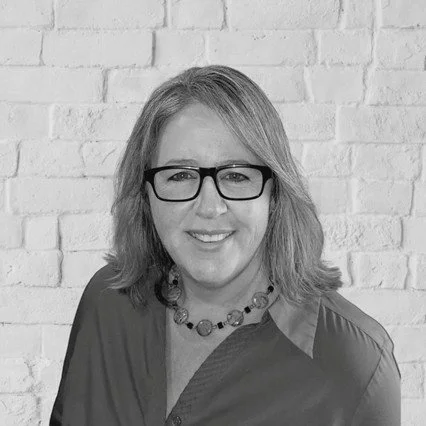 A woman with light hair and glasses wears a collared blouse.