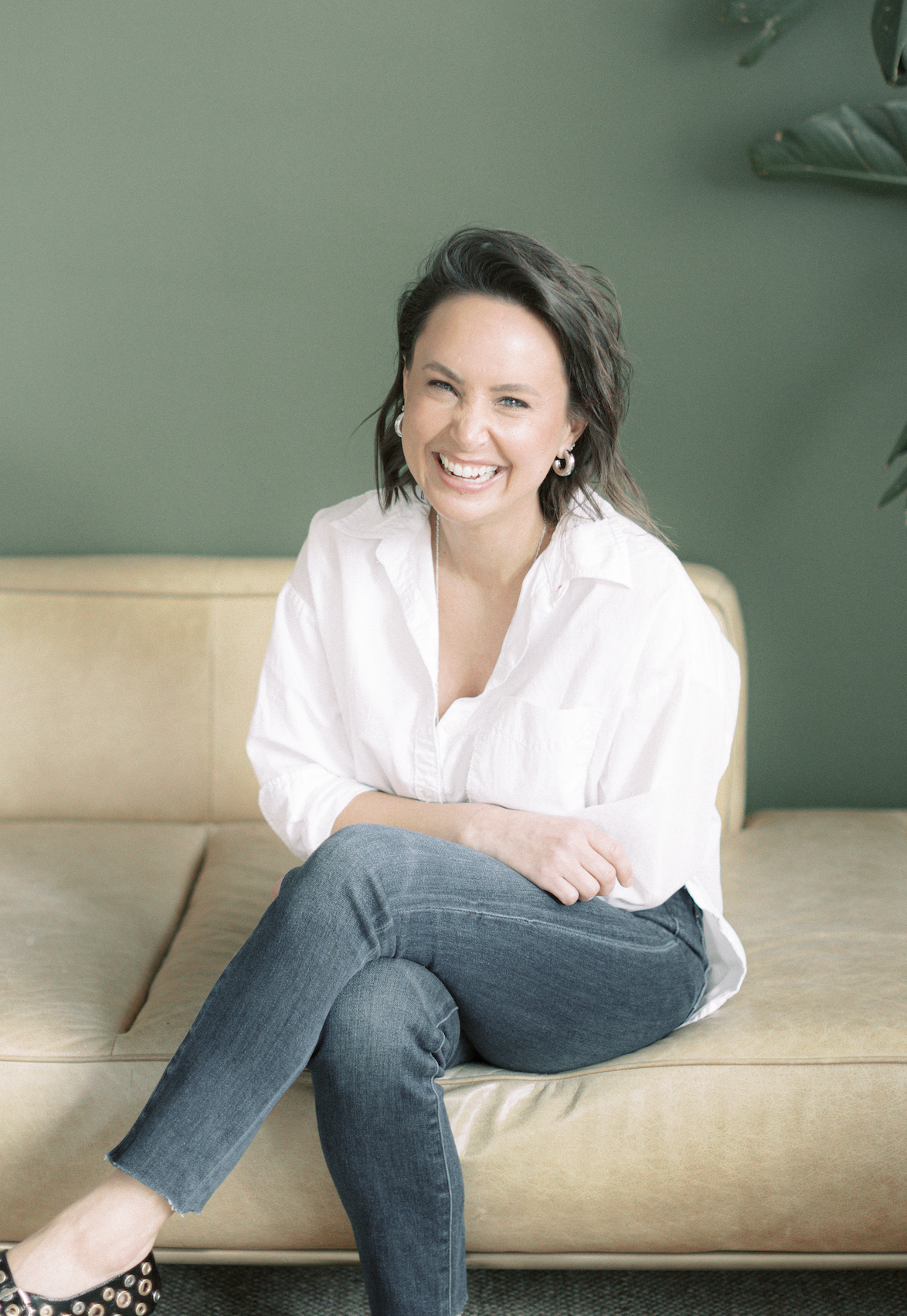 A woman wearing a crisp white blouse and jeans sits on a beige sofa, smiling