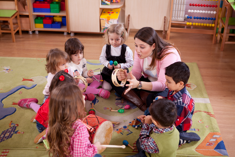 An early childhood caregiver or teacher leads children in a music group. Children sit in a cirlce and hold percussion instruments.