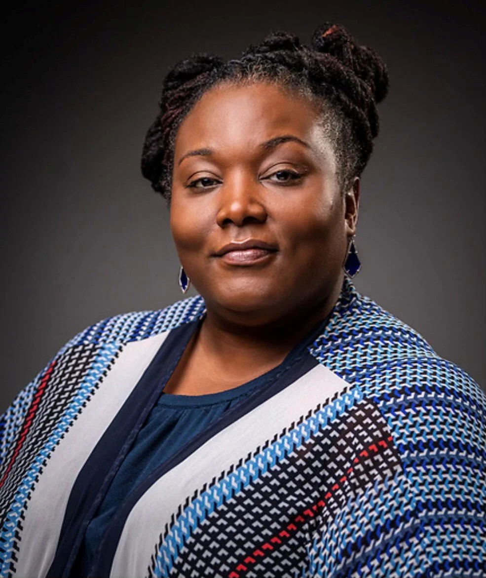A woman with dark hair coiled on her head wears a brightly colored top.