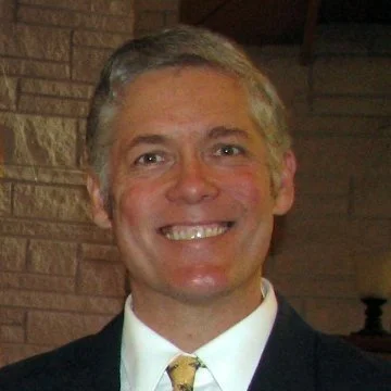 A man with grey hair wears a suit and stands in front of a stone wall backdrop