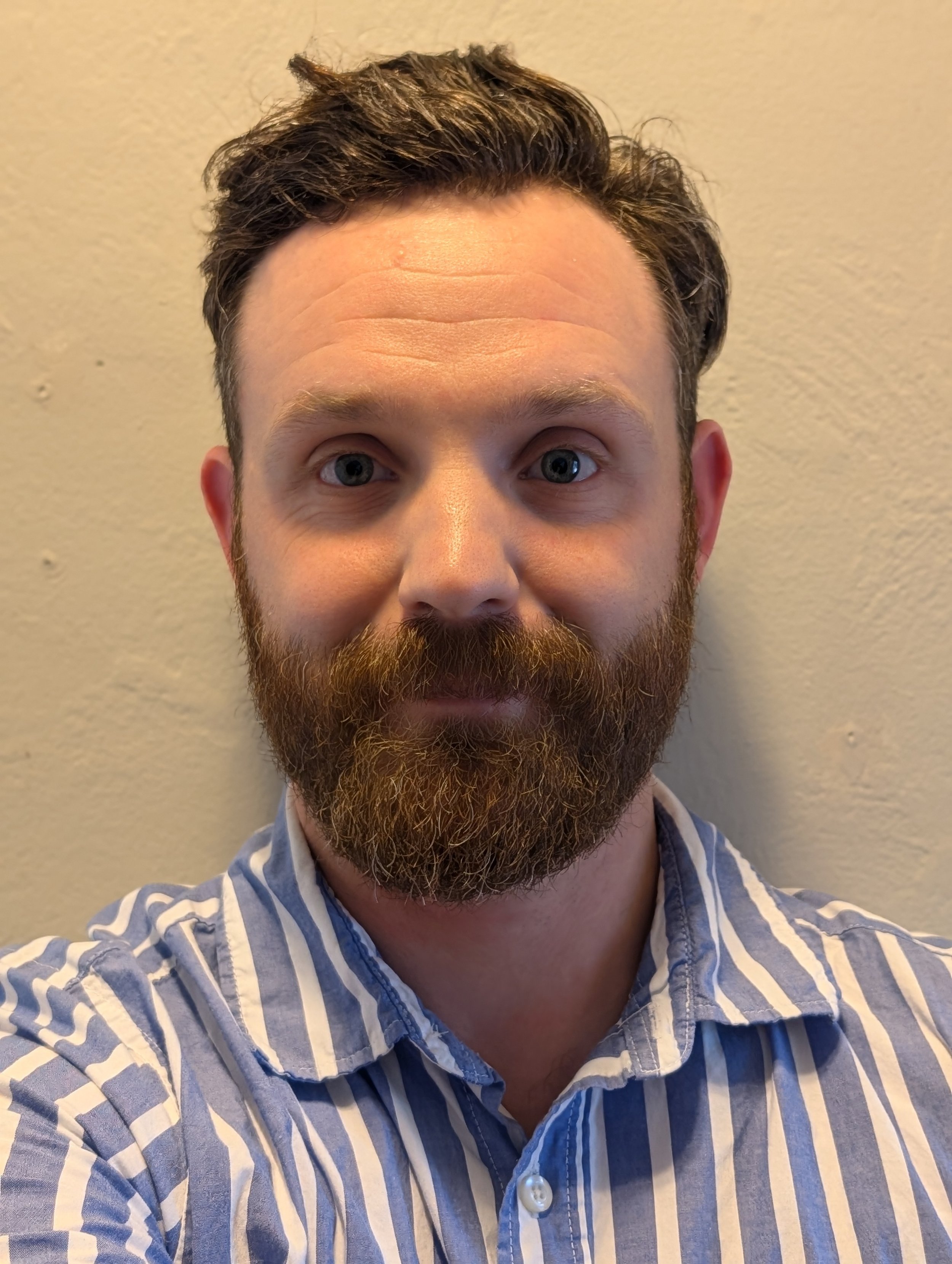 Jeremy is shown with a trimmed beard and mustache, wearing a striped blue and white shirt