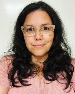 A woman with long, dark, wavy hair wears pale, cat-eye style glasses and a pink blouse, looking directly at the camera against a light background.