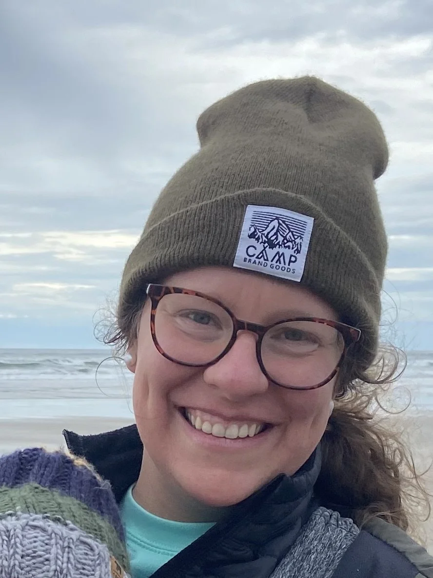 a smiling woman in a beanie and glasses at a beach, with the ocean in the background.