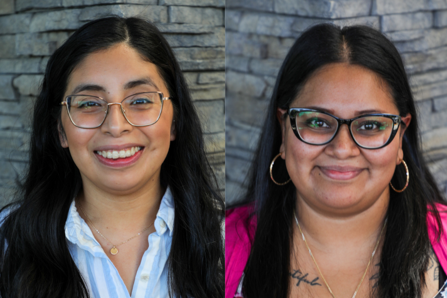 Britania and Maria are women with long brown hair and glasses. In this image, they smile in front of a stone wall