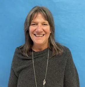 A woman with light brown and gray shoulder-length hair wears a gray sweater and a long chain necklace,