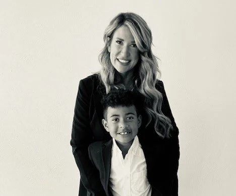 A black-and-white portrait of a smiling woman with long wavy hair standing behind a young boy in a blazer and white shirt; both are looking directly at the camera.