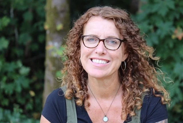 woman with long curly red hair smiles in front of trees