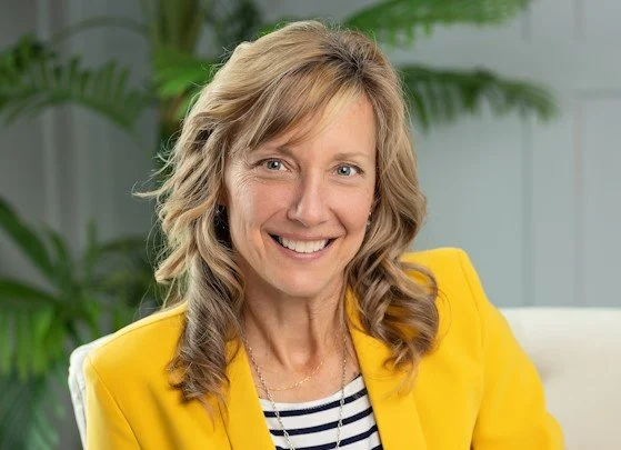 Diane is pictured with wavy light brown hair, light eyes, and wearing a bright yellow blazer