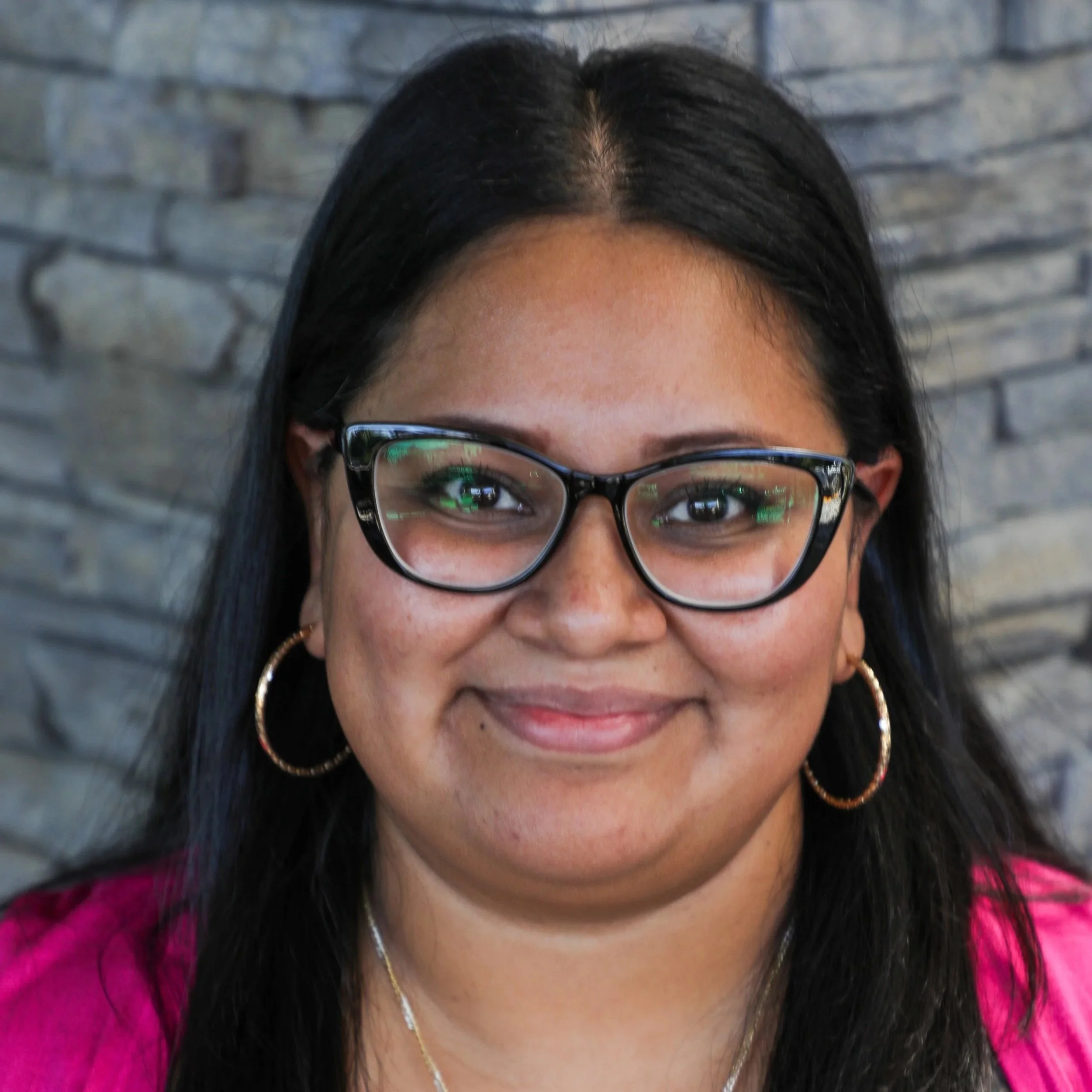 Maria is shown with dark eyes behind bold framed glasses, long dark hair, and wearing a pink top